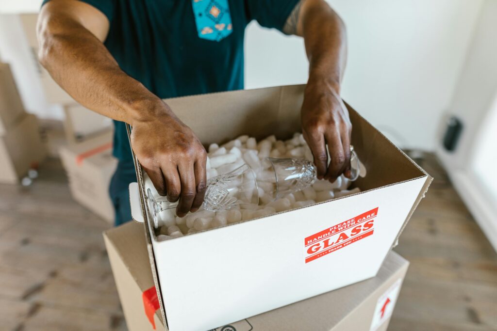 hands carefully packing glassware into a cardboard box with padding for safe transport.