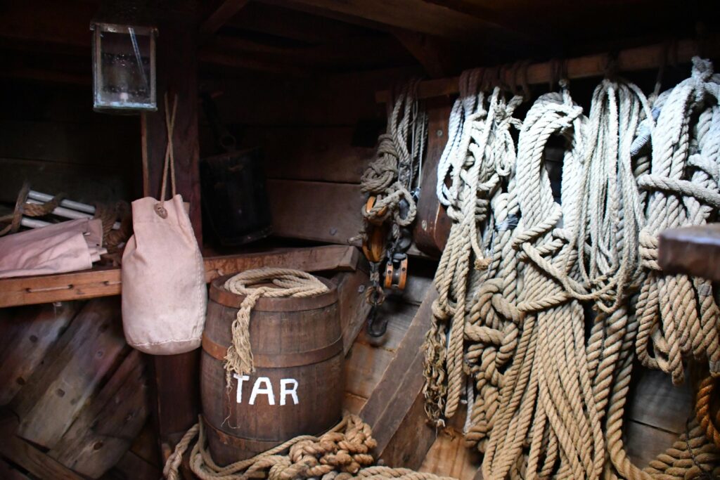 vintage ship interior showing a wooden barrel labeled 'tar' surrounded by coiled ropes and rustic decor.