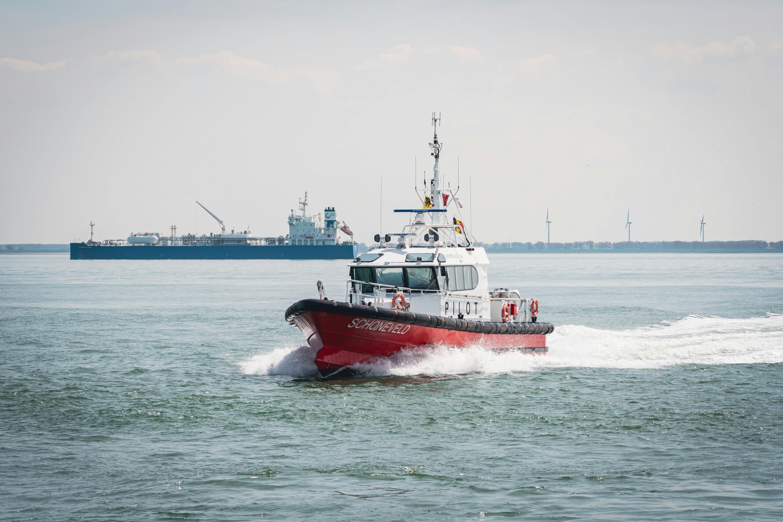 a red pilot boat travels across the sea with ships in the background and wind turbines on the horizon.