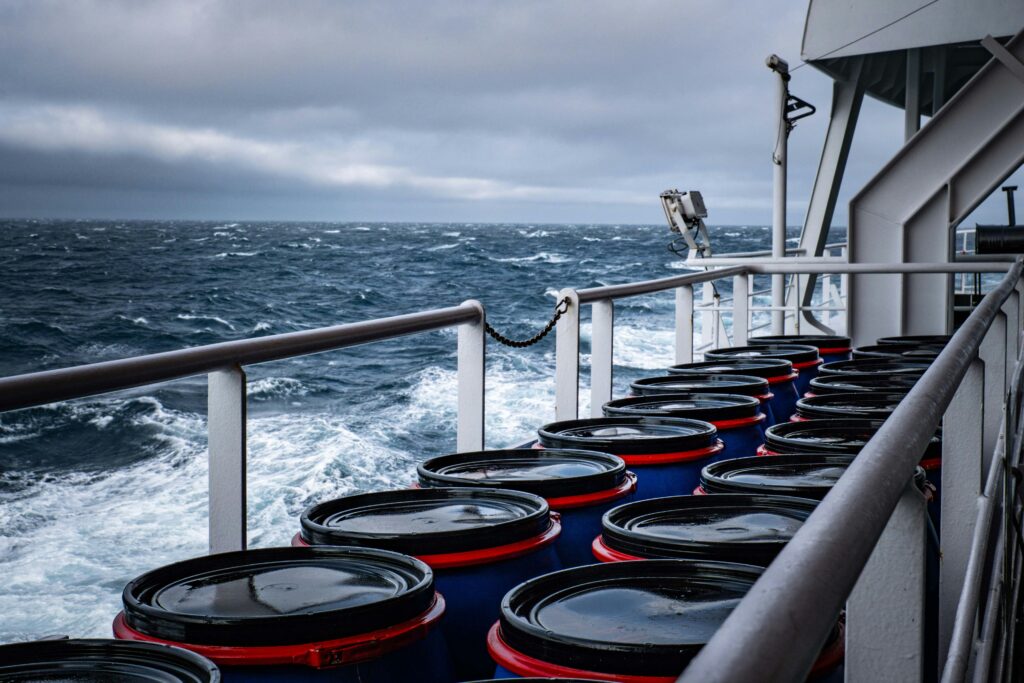 industrial barrels stacked on a ship deck amidst a stormy sea, suggesting maritime shipping.