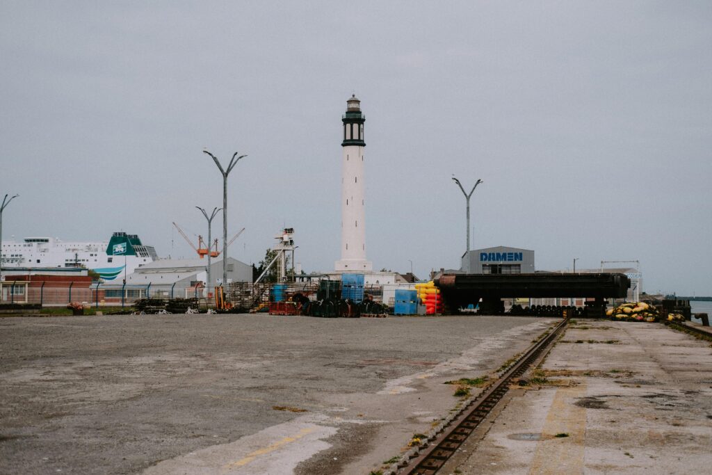 a lighthouse stands tall in an industrial port setting with a ferry in the background.