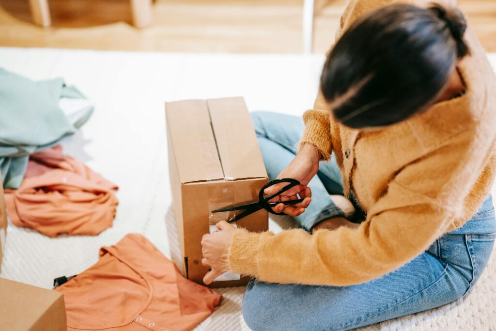 woman carefully packs clothes into a box with scissors for shipment indoors.